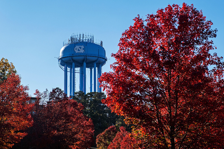 Water tower with U.N.C. logo in behind trees with red leaves.