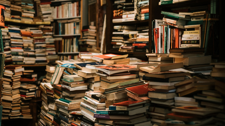 A photo of a pile of books on a table and shelves stuffed with books.