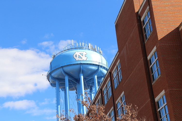A photo partially showing the ITS Manning building (which is red brick) with a water tower in the background. The water tower is Carolina blue and has a white interlocking NC emblem painted on it. 