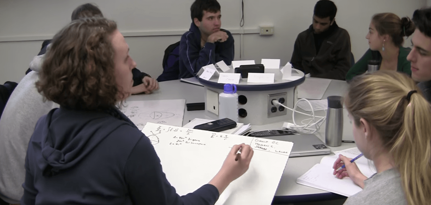 Students sit around a table while a few write on large poster board