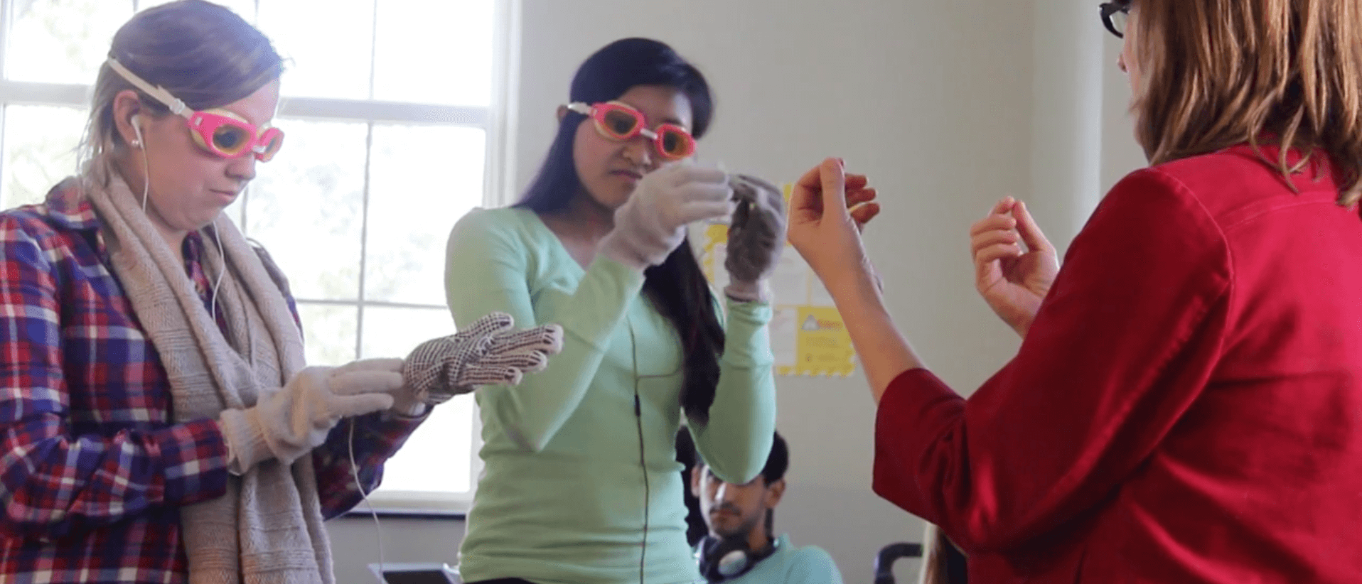Two students in a classroom wear googles and earbuds and put on gloves as their professor talks to them.