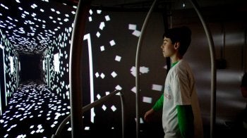 A middle school students stands in he dark surrounded by square lights.