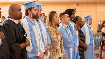 Students in caps and gowns and faculty members standing and placing their hands over their hearts.