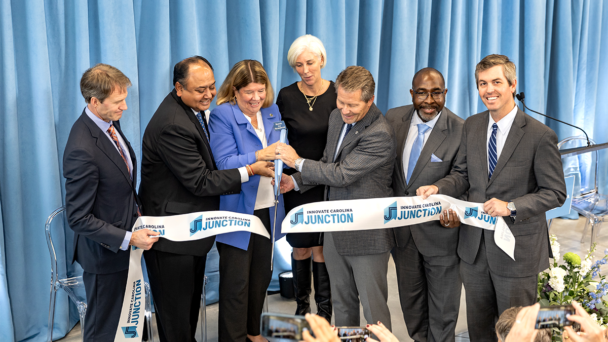 A group of six people standing in front of an audience and cutting a ribbon with a large pair of scissors at a ribbon-cutting event for the Innovate Carolina Junction.