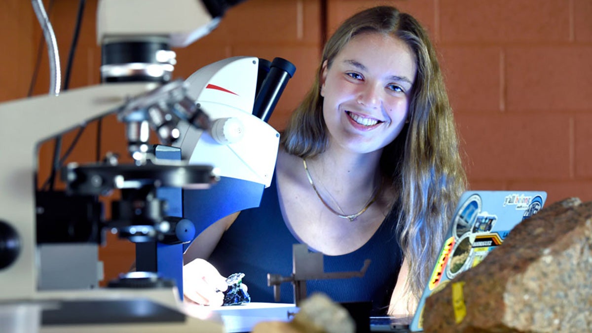 Olivia Key pokes her head from behind a microscope in the foreground.