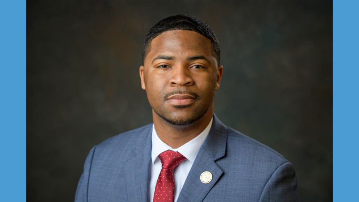 Headshot of Gilbert Rogers wearing suit jacket and red tie against a grey backdrop, surrounded by a Carolina Blue border.