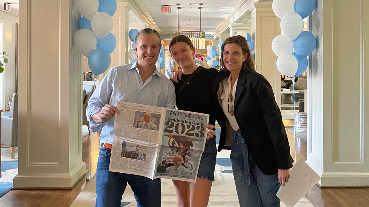 Carolina Ramirez and her parents posing with open newspaper in front of hallway adorned with Carolina Blue balloons.