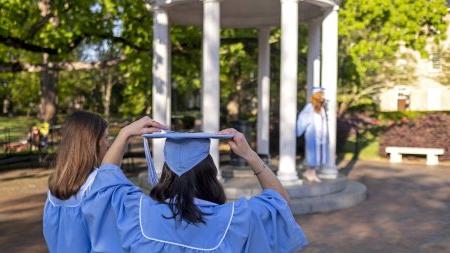A student wearing a cap and gown with her back to the camera. She's waiting by the Old Well as another student in regalia has a graduation photo taken at the Old Well.