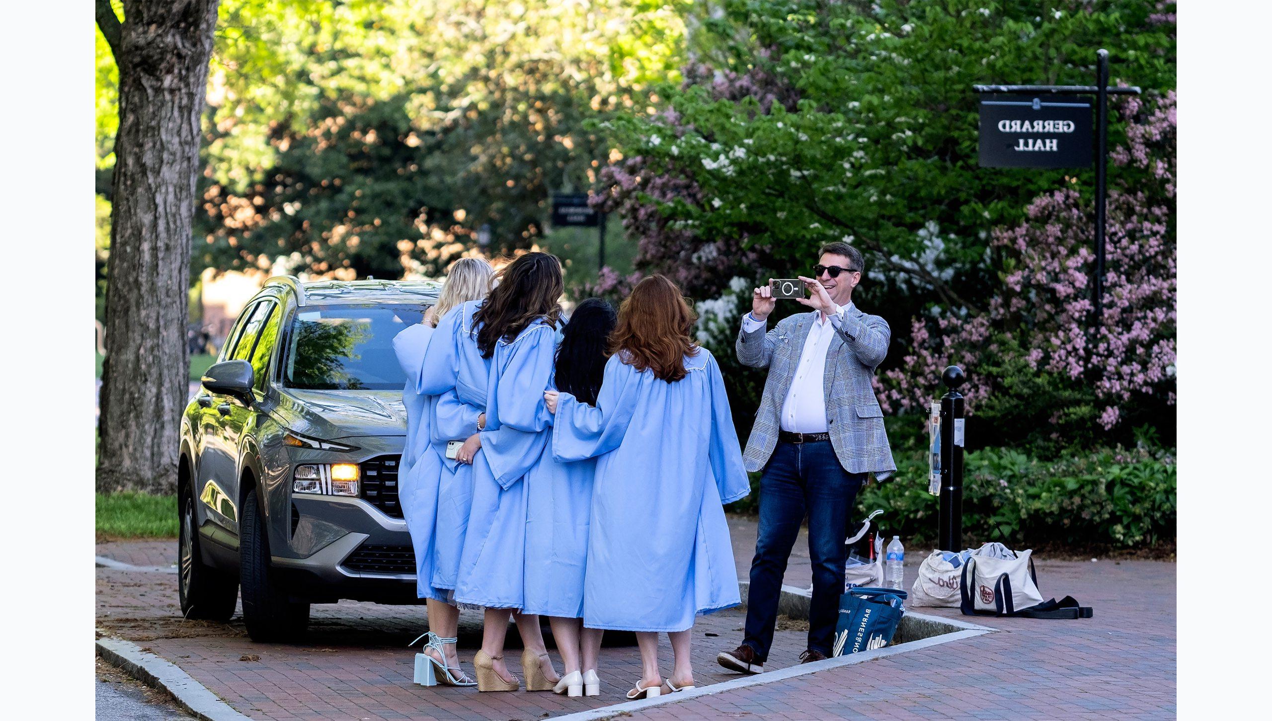 A man taking a picture with a phone of a group of students in graduation gowns.