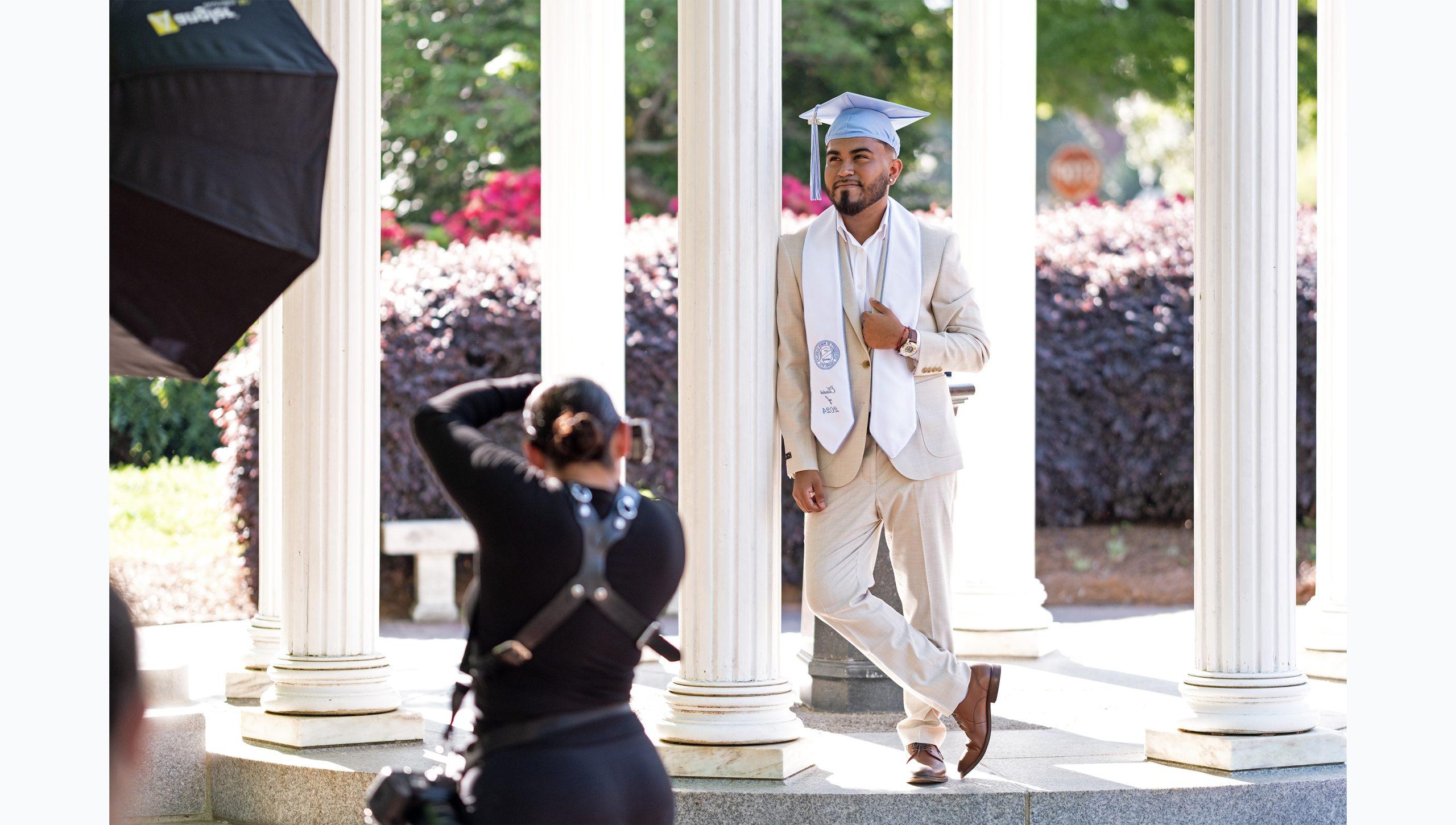 A student posing for a graduation photo at the Old Well in a suit with his graduation cap.