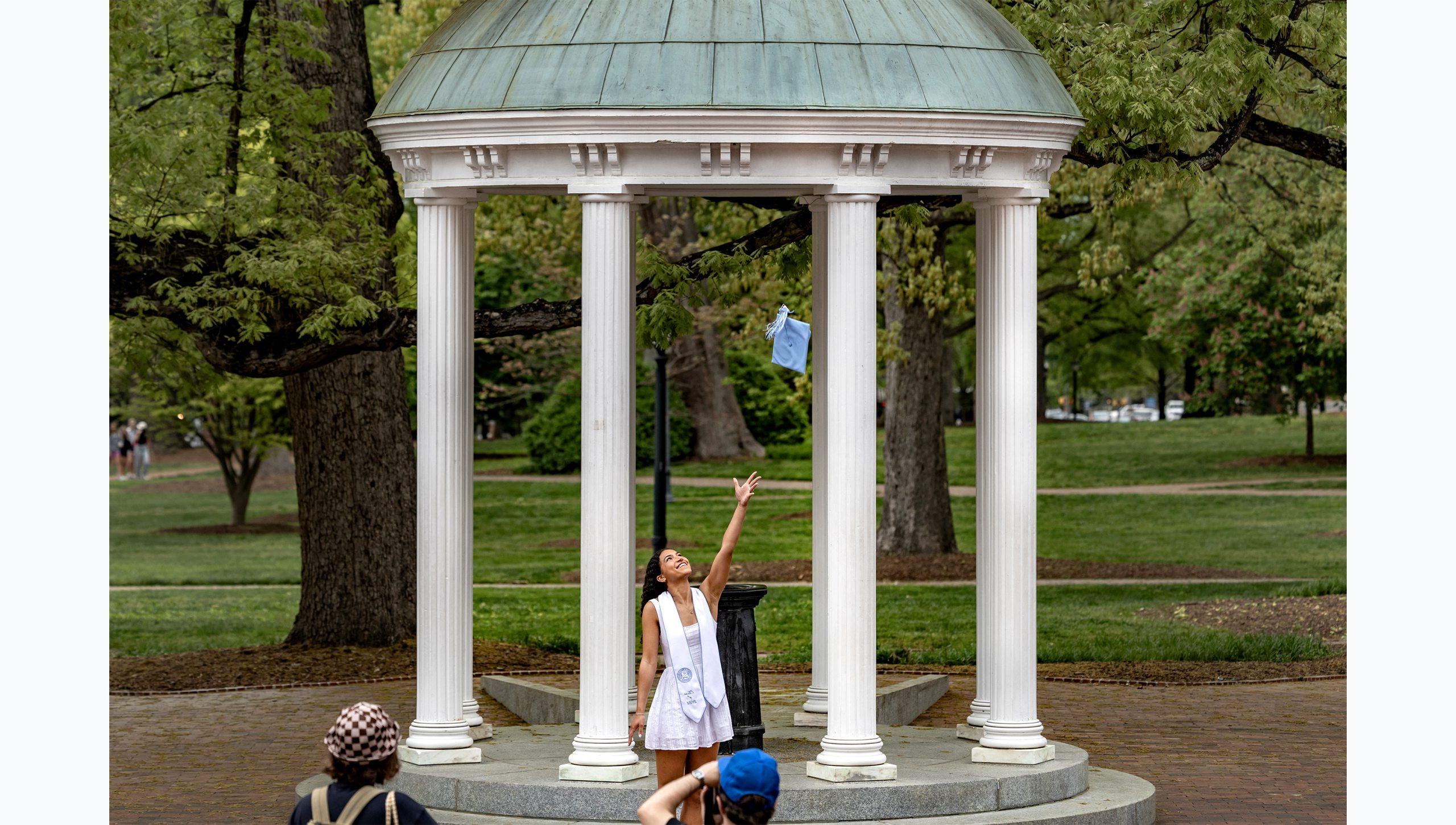 A student tossing her cap up in the air as she poses for a graduation photo at the Old Well.