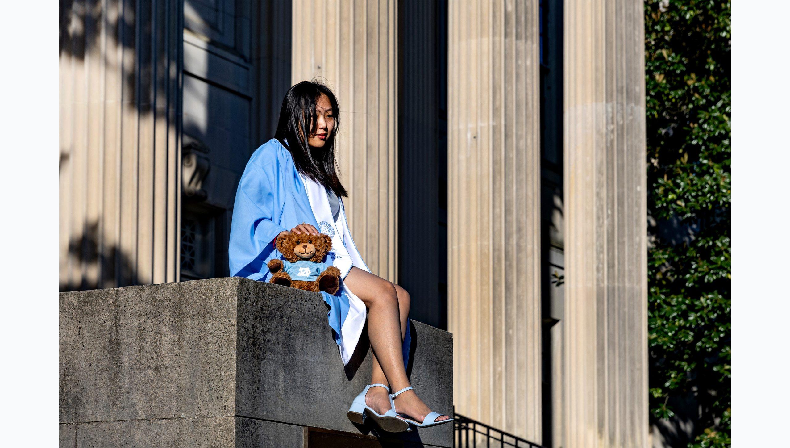 A student posing for a graduation photo with a UNC Teddy Bear in front of Wilson Library.