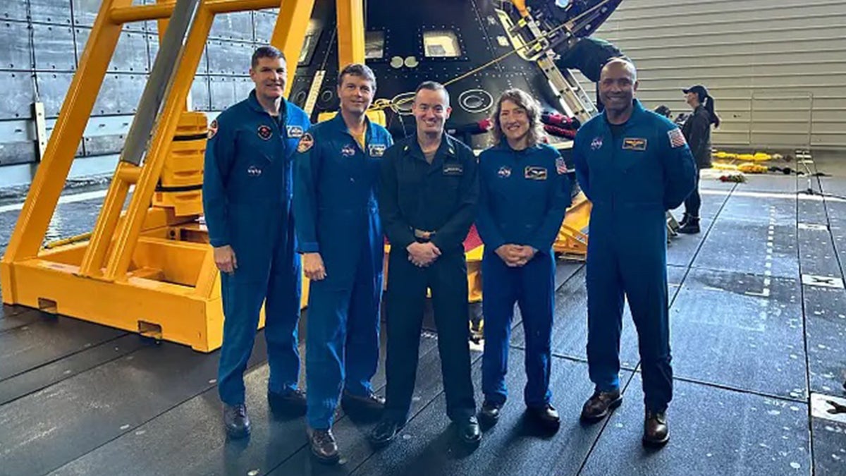 Five individuals posing in a row wearing blue NASA flight suits.