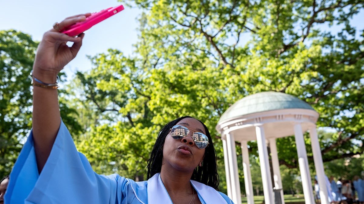 Student in graduation gown takes selfie in front of Old Well