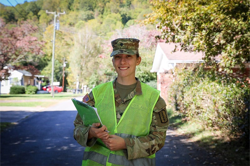 Jessica Bowling in National Guard uniform holding a clipboard and smiling for a portrait while standing on a street in a neighborhood in Waynesville, North Carolina.