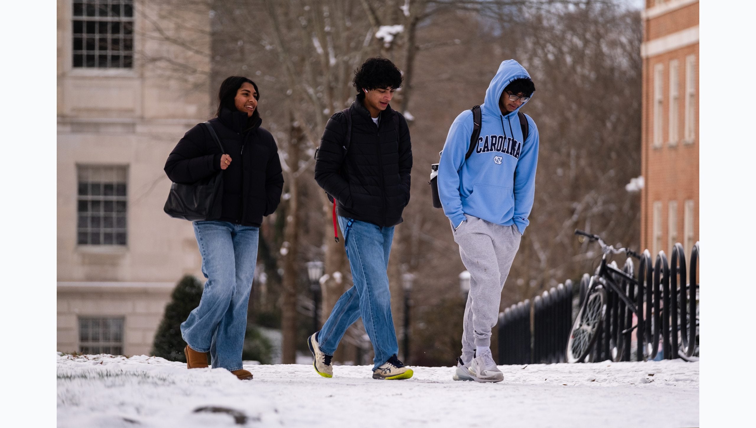 Three students walking along snow-covered pathway on the campus of U.N.C Chapel Hill. One of the three students is wearing a Carolina Blue Tar Heels hoodie.