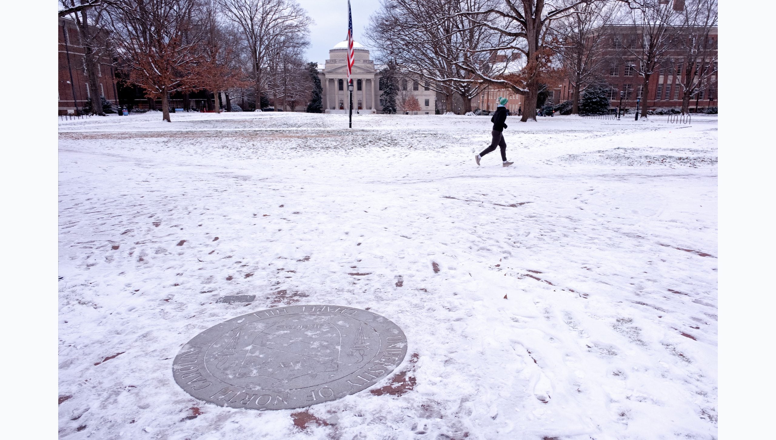 The University seal cleared and displayed among an otherwise snow-covered Polk Place on the campus of U.N.C Chapel Hill campus. A runner is seen in the background.