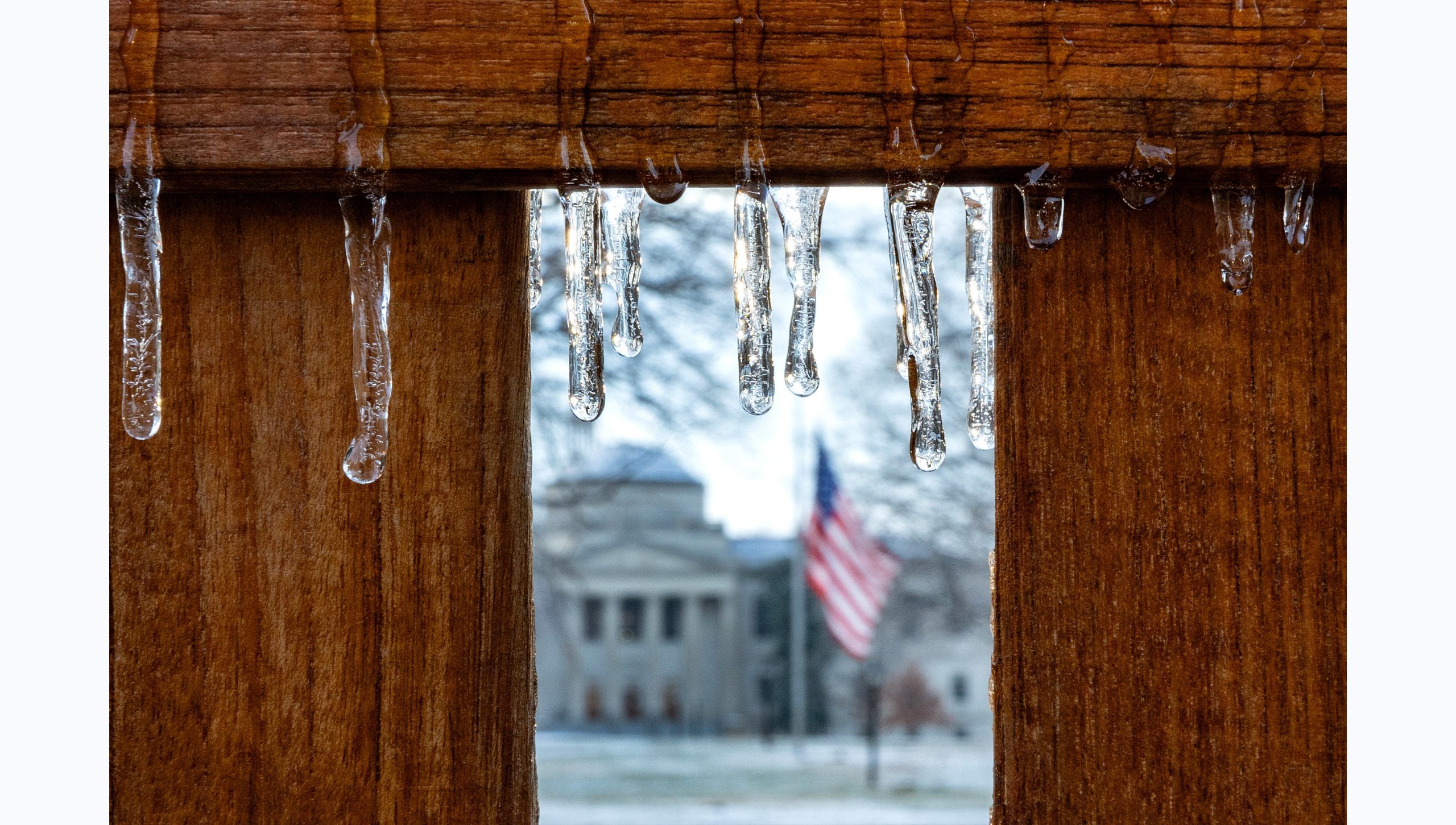 Close-up image of icicles hanging off a wooden bench on the campus of U.N.C Chapel Hill campus. Seen in the background is an American Flag and Wilson Library.