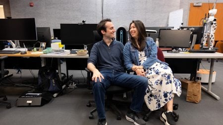 Two UNC-Chapel Hill graduate students who are engaged to each other, Angelos Angelopoulos and Jade Kandel, sit and hold hands in a laboratory with computer screens seen behind them.