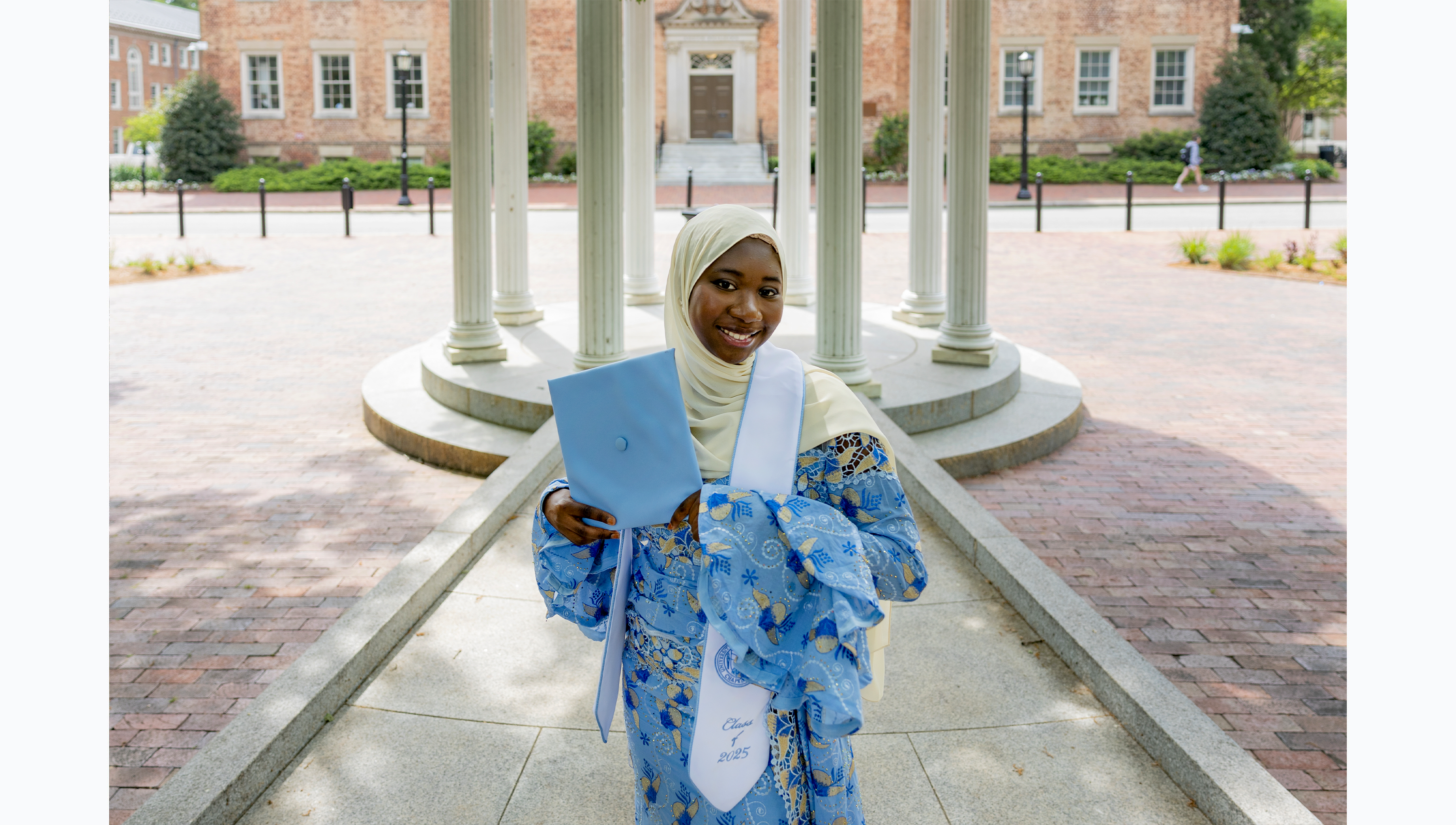 A student in Carolina Blue and white regalia holding up her cap while posing for a graduation photo at the Old Well.