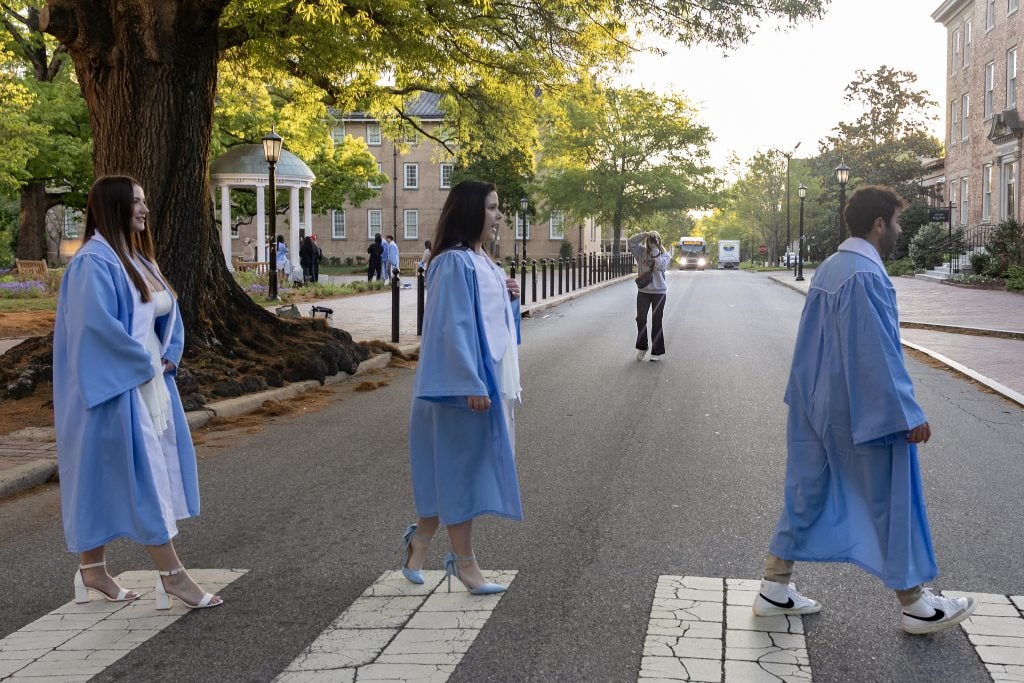 Shaena Riddles, Brittany Harrelson and Nick Clementi in their Carolina Blue caps and gowns crossing East Cameron Avenue in front of the Old Well as Samantha Lewis, in the background, makes a picture of them.