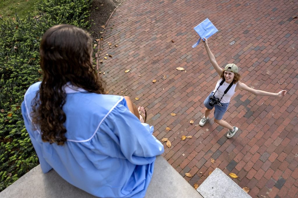 Photographer Heather Diehl ’25 extending her arms wide to direct Riley Quartermain ’25 on what to do while making graduation photos for her near the steps of Wilson Library on the campus of UNC-Chapel Hill.