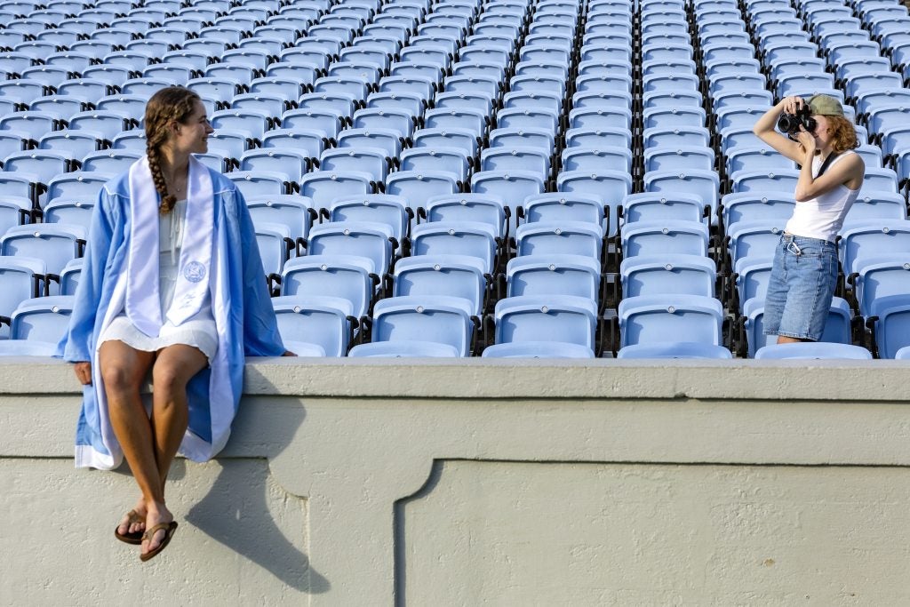 UNC-Chapel Hill Riley Quartermain sitting on a ledge in front of rows of seats at Kenan Stadium as Heather Diehl, several feet over to the right, makes a picture of her.