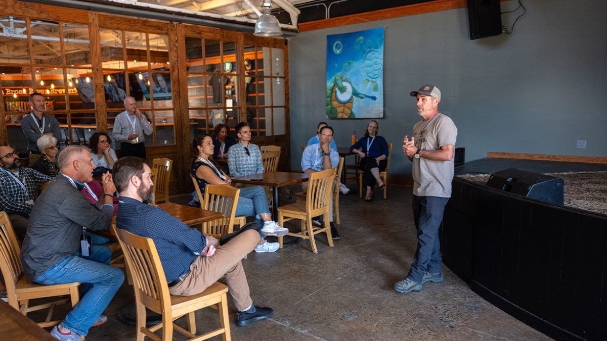 Brewery owner speaking to seated Bus Tour attendees in his brewery.