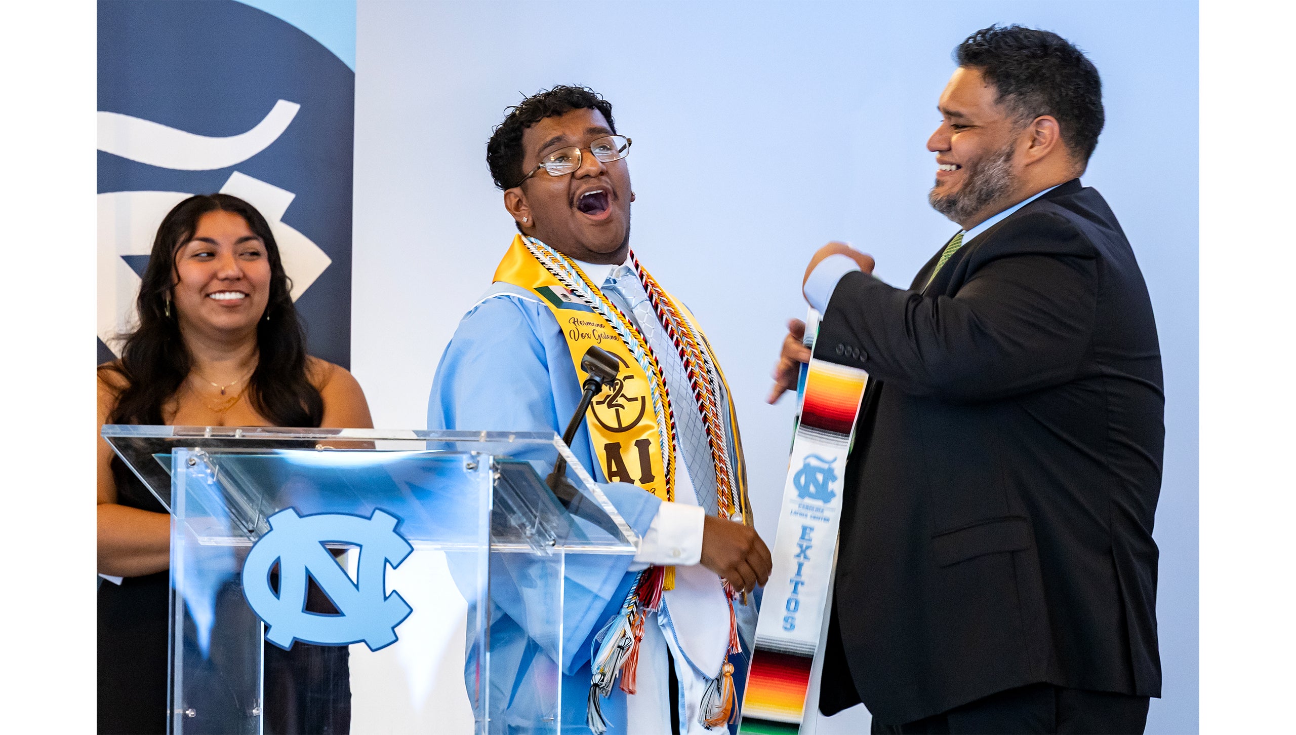 A graduate smiles on stage next to classmates at the Exitos Award Ceremony