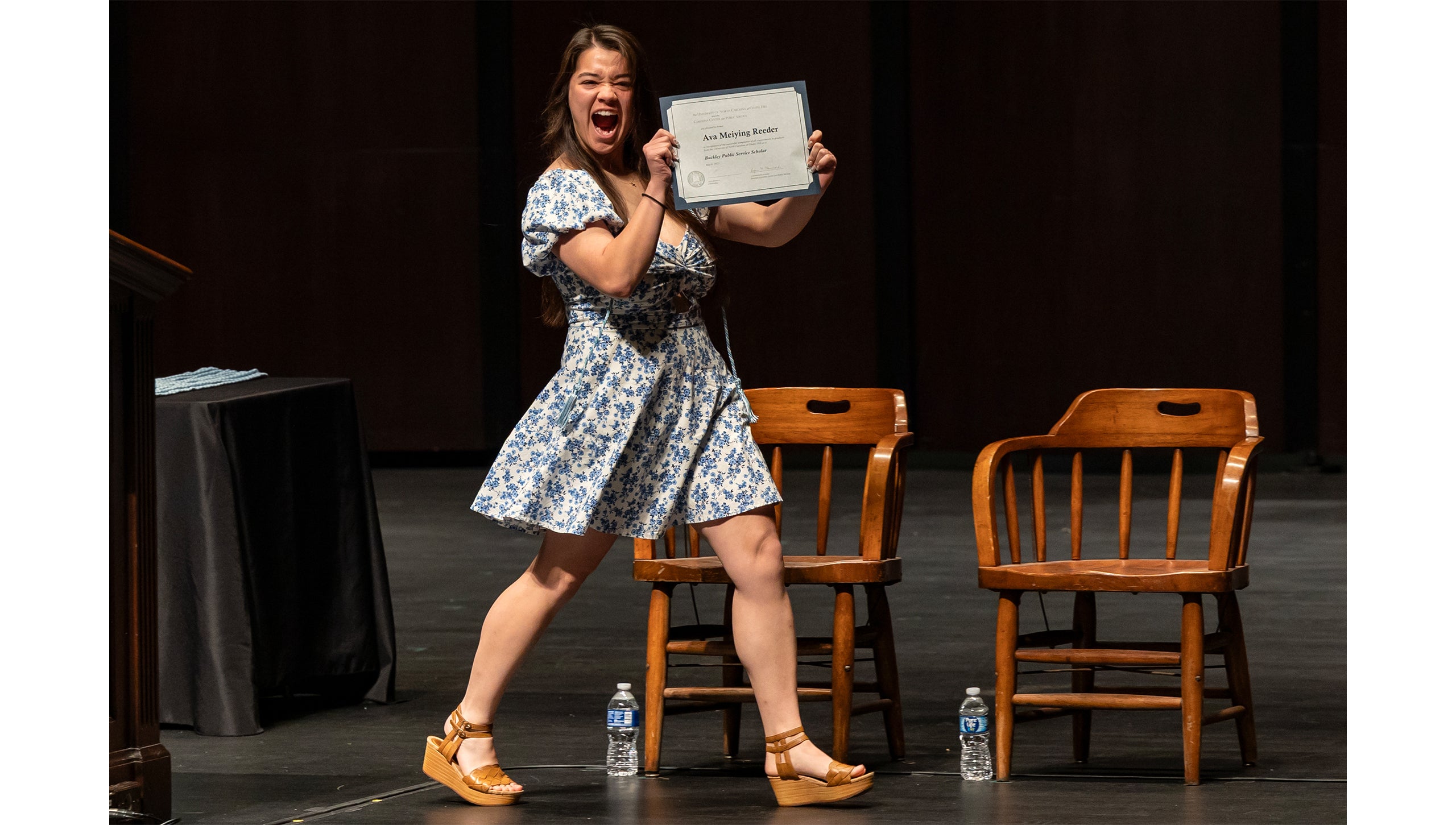 A graduate smiles while holding a certificate and crossing the stage at the Buckley Public Service Scholars Ceremony