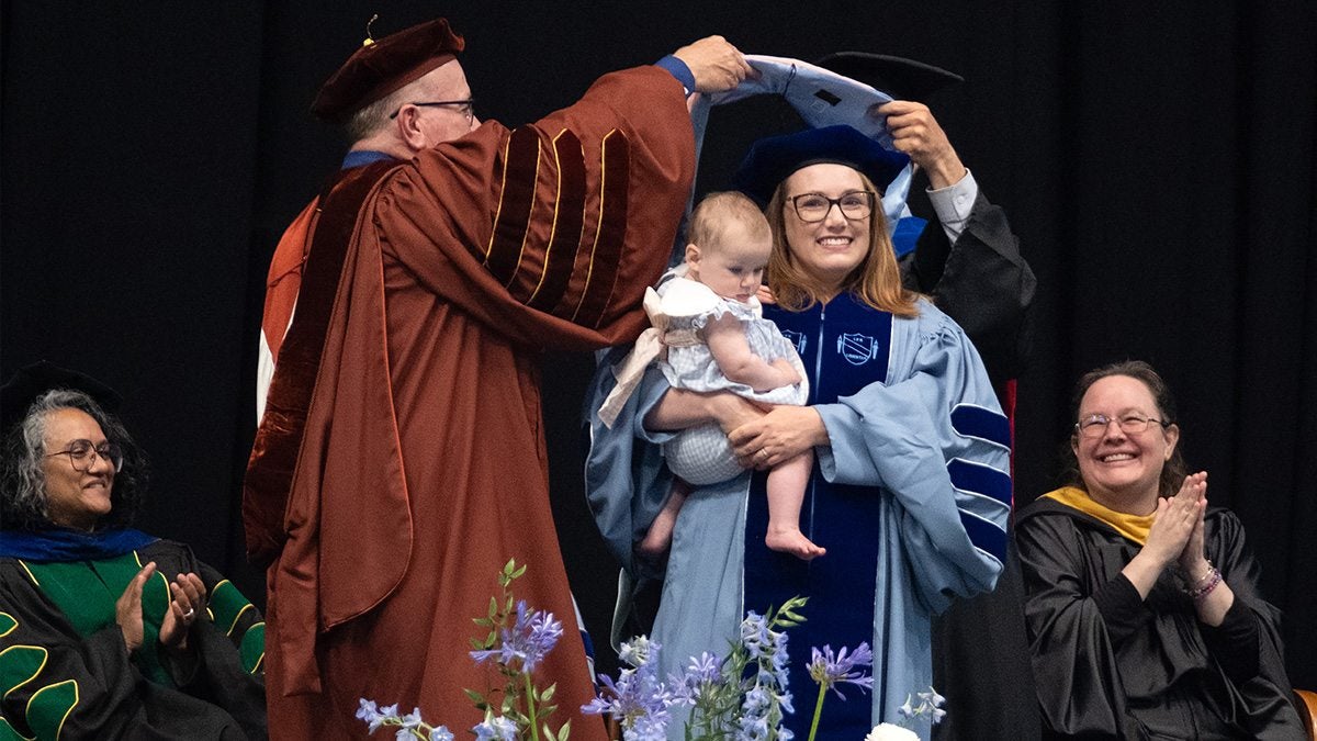 A doctoral graduate is hooded while holding her baby on stage at the doctoral hooding ceremony
