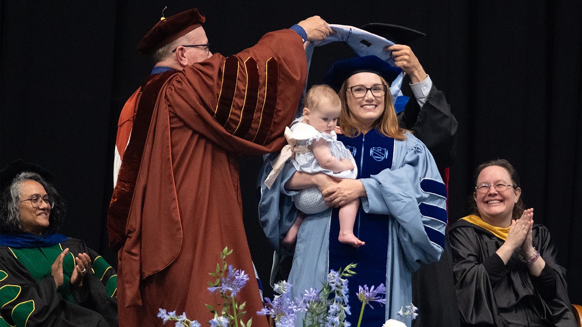 A doctoral graduate is hooded while holding her baby on stage at the doctoral hooding ceremony