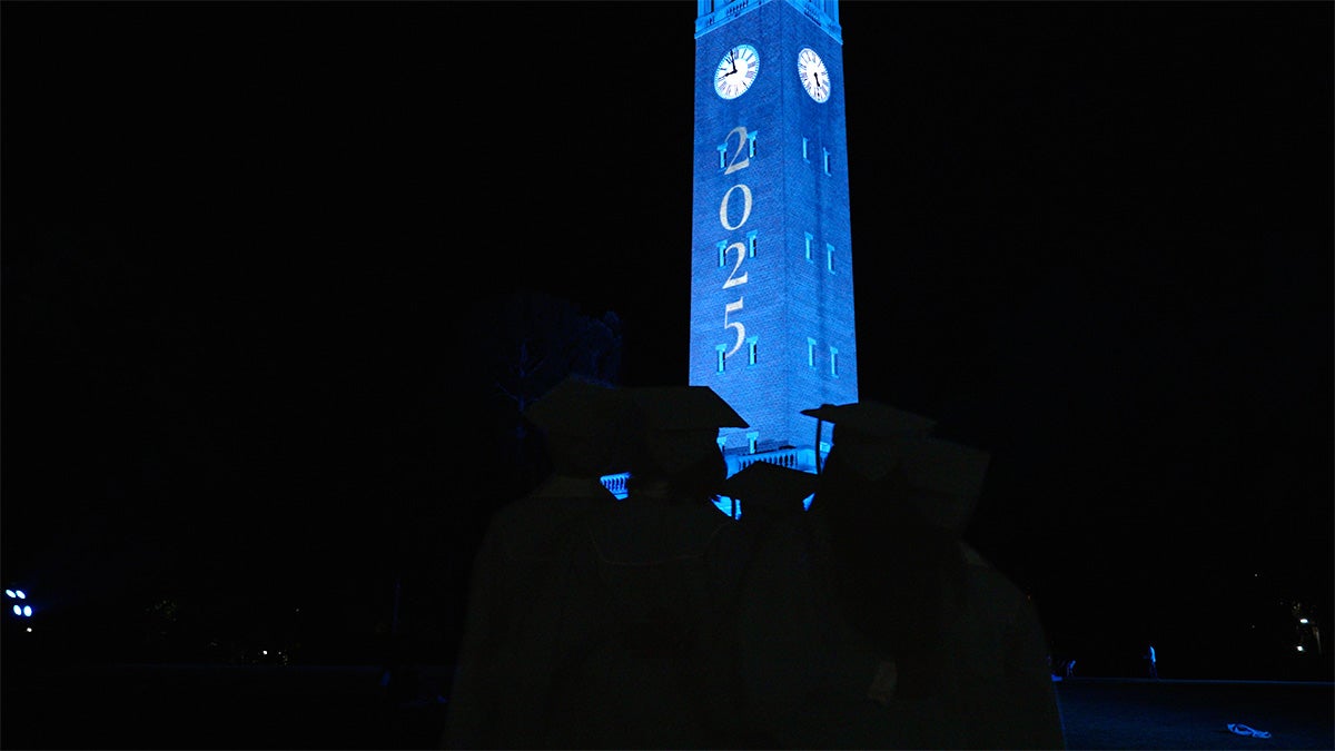 UNC-Chapel Hill's Morehead-Patterson Bell Tower lit Carolina Blue in the dark the night of Commencement and projected white text reading 