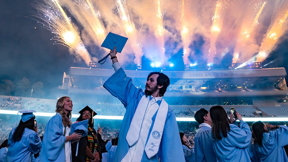 A graduate holds his cap in the air as fireworks go off behind him at Kenan Stadium during 2025 Spring Commencement.