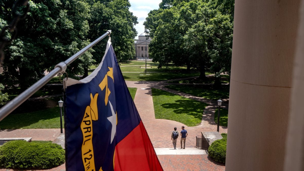 State of North Carolina flag flying from South Building on the campus of UNC-Chapel Hill. Polk Place, with two people pictured, is seen in the background.