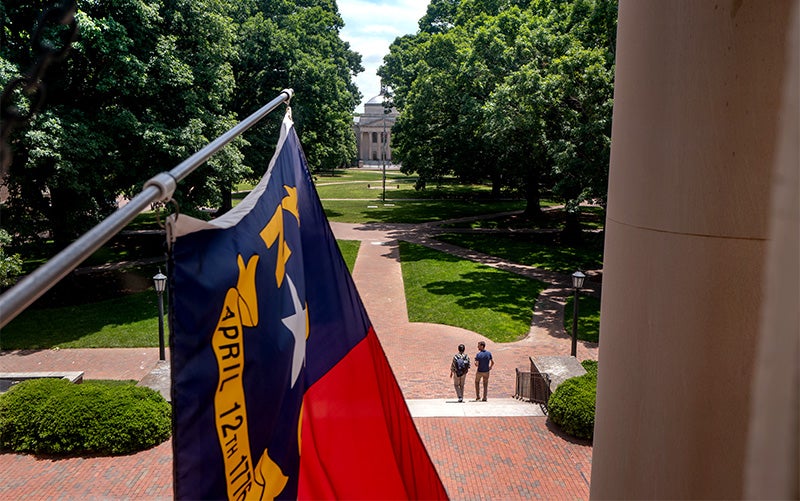 State of North Carolina flag flying from South Building on the campus of UNC-Chapel Hill. Polk Place, with two people pictured, is seen in the background.