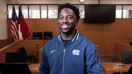 Charles Hands posing for portrait and wearing a Navy blue UNC jacket in a court room.