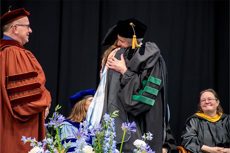 Kim Enders and her father, Dr. Bert Peterson, hugging onstage at the doctoral hooding. Others smile as they watch the scene.