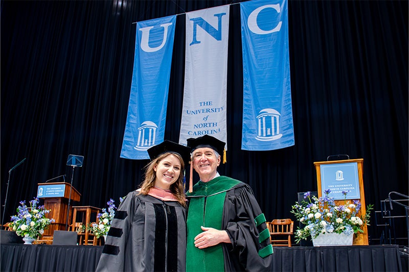 Kim Enders and her father, Bert Peterson, posing for photo in black regalia at UNC-Chapel Hill's doctoral hooding in front of the stage. "UNC" banners are seen hanging in the background.