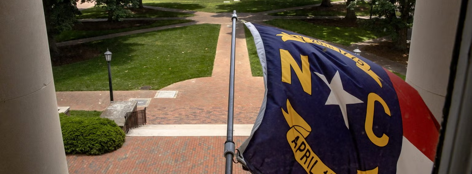 North Carolina state flag flying from the the exterior of South Building on the campus of UNC-Chapel Hill. Polk Place, known colloquially as the Quad, is seen in the background.
