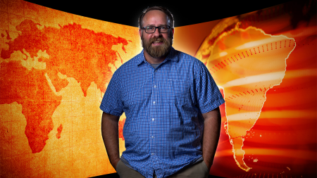 Clark Gray posing for a photo. In the back is a graphic design of large global maps colored orange.