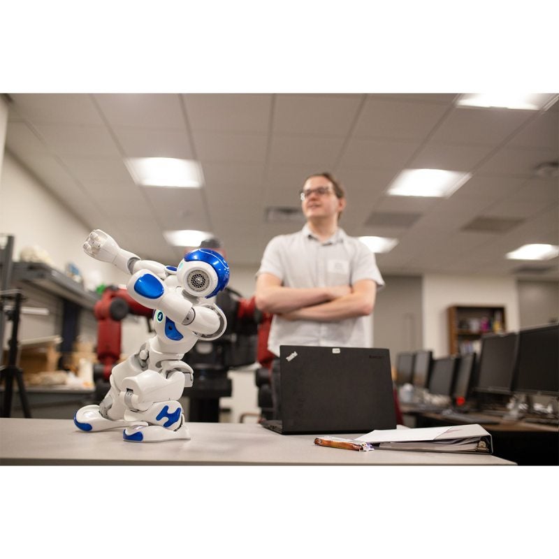 A miniature robot toy on a desk. A person with a laptop is standing in the background.