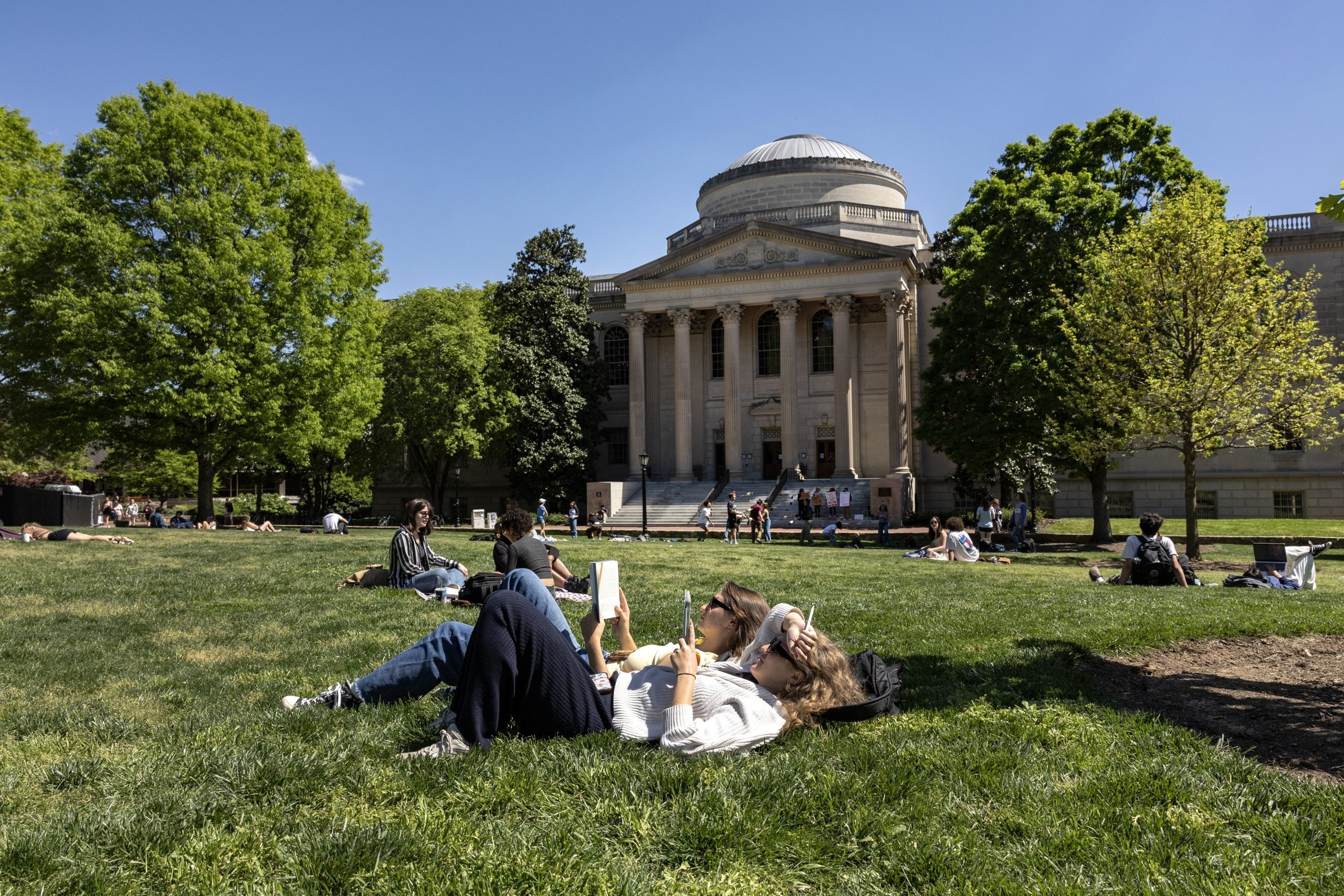 Two students lying down and reading on the grass of Polk Place on the campus of UNC-Chapel Hill with Wilson Library seen in the background.