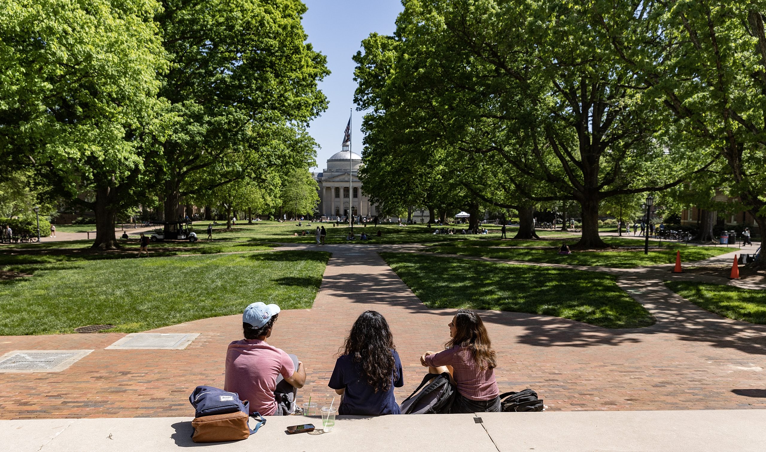 Three students sitting on the steps of South Building facing Wilson Library and Polk Place on the campus of UNC-Chapel Hill.