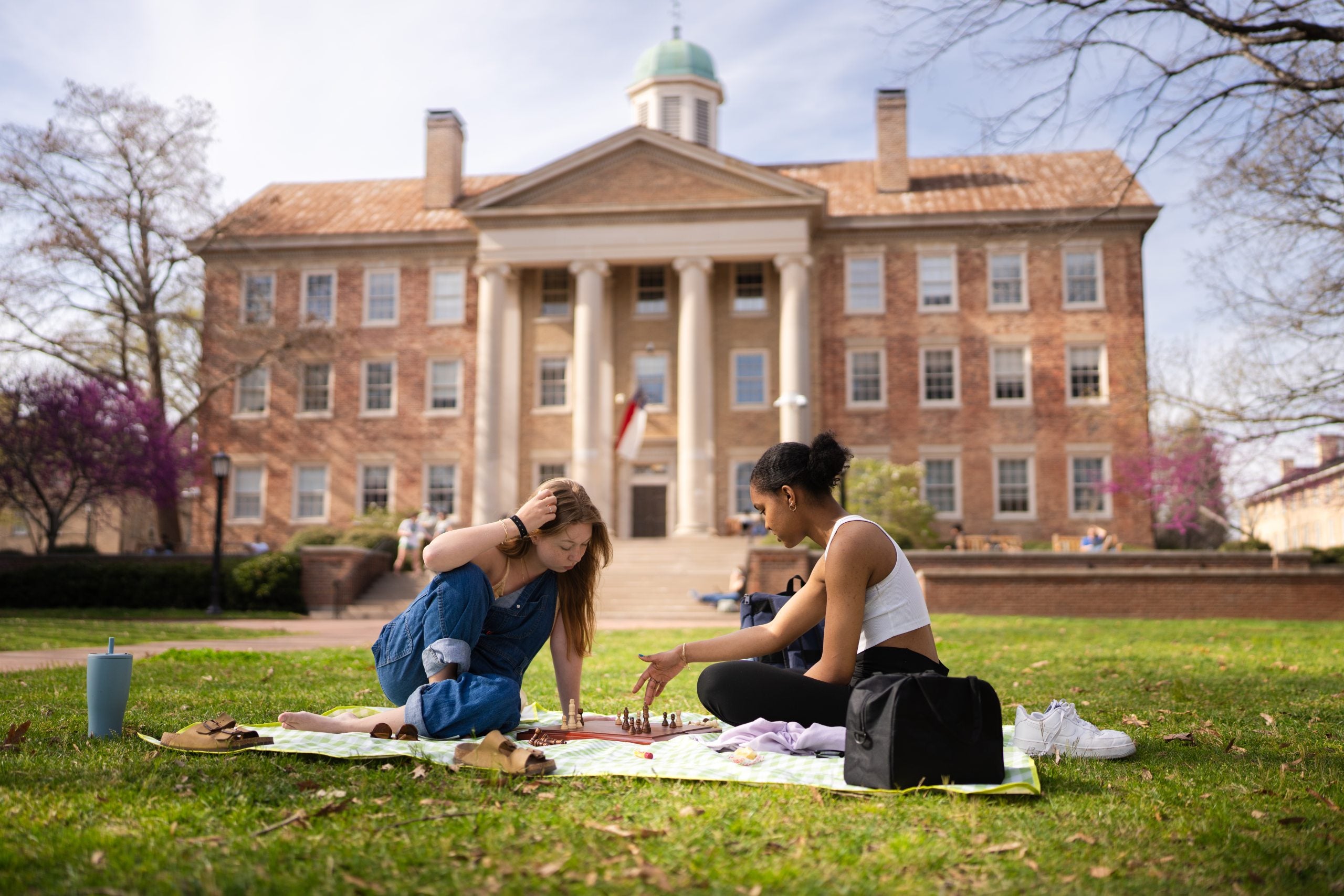 Two female students sitting on a towel and playing chess on the lawn of Polk Place on the campus of UNC-Chapel Hill. South Building is seen in the background.