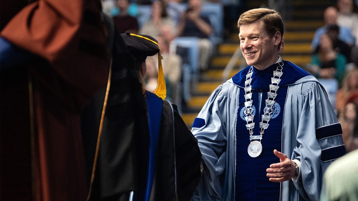 Chancellor Lee H. Roberts smiling and shaking someone's hand while wearing Carolina Blue regalia with a medallion on at UNC-Chapel Hill's doctoral hooding inside the Dean E. Smith Center.